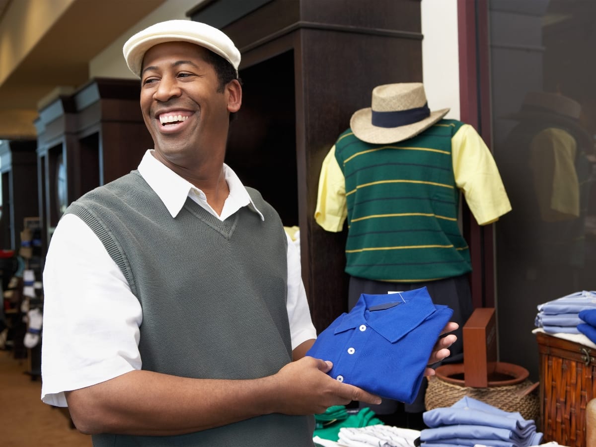 A man wearing a flat cap and vest smiles while holding a folded blue polo shirt in a clothing store, showcasing Atlanta Golf Apparel Trends among other stylish shirts and mannequins in the background.