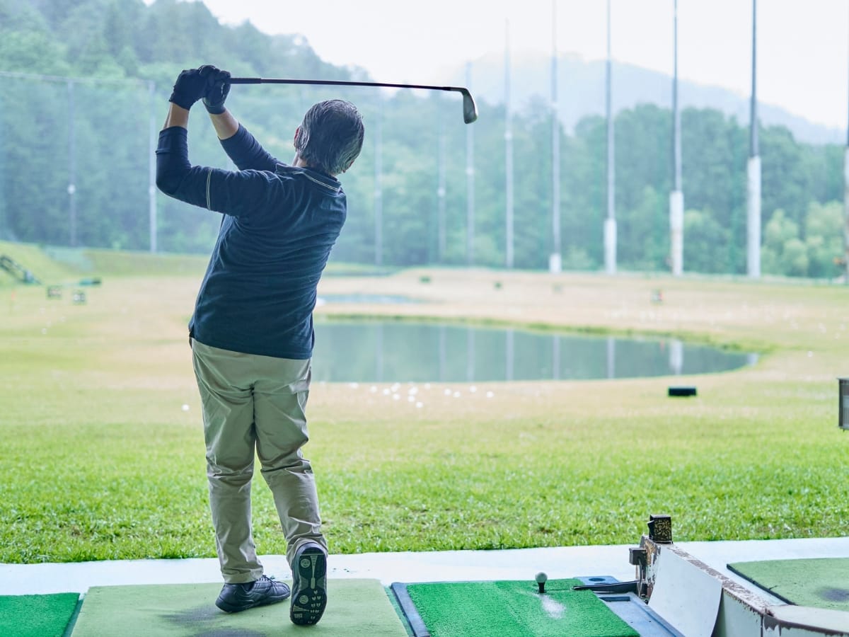 A person practices golf at a driving range, swinging a club toward an open grassy field with a pond and trees in the background, dressed in the best golf apparel for Atlanta weather.
