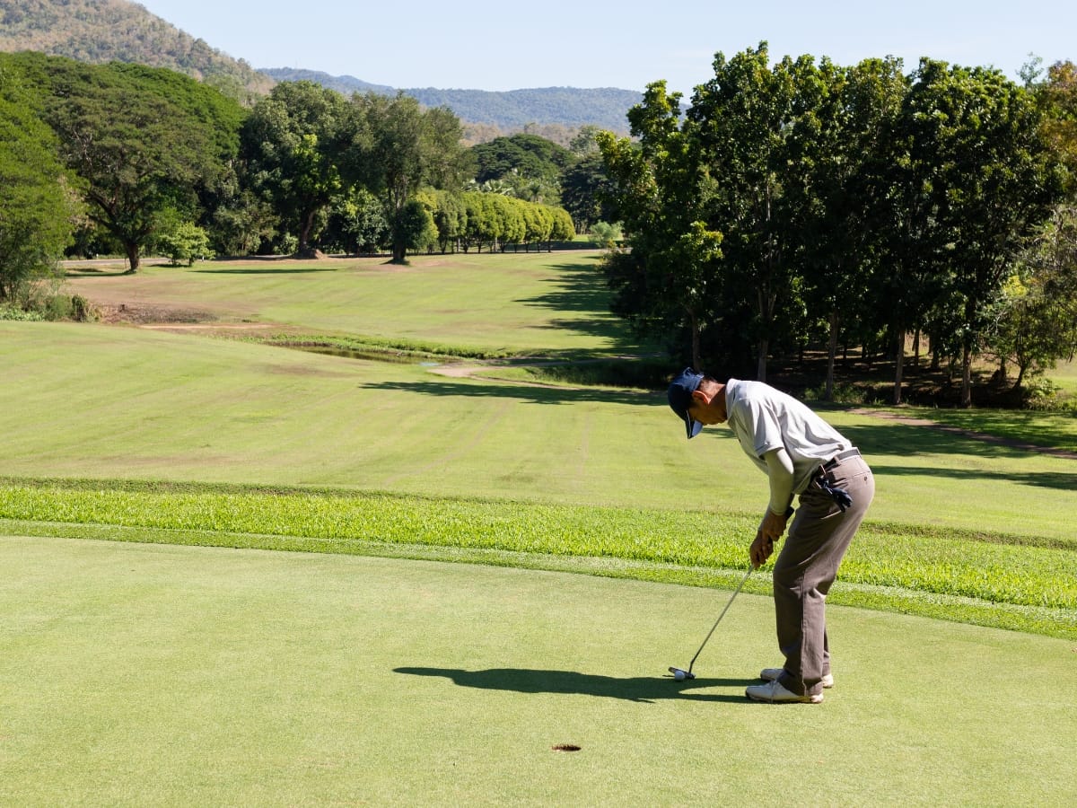 A golfer in a white shirt and cap, sporting the best golf apparel for Atlanta weather, putts on a green with trees and hills in the background on a sunny day.