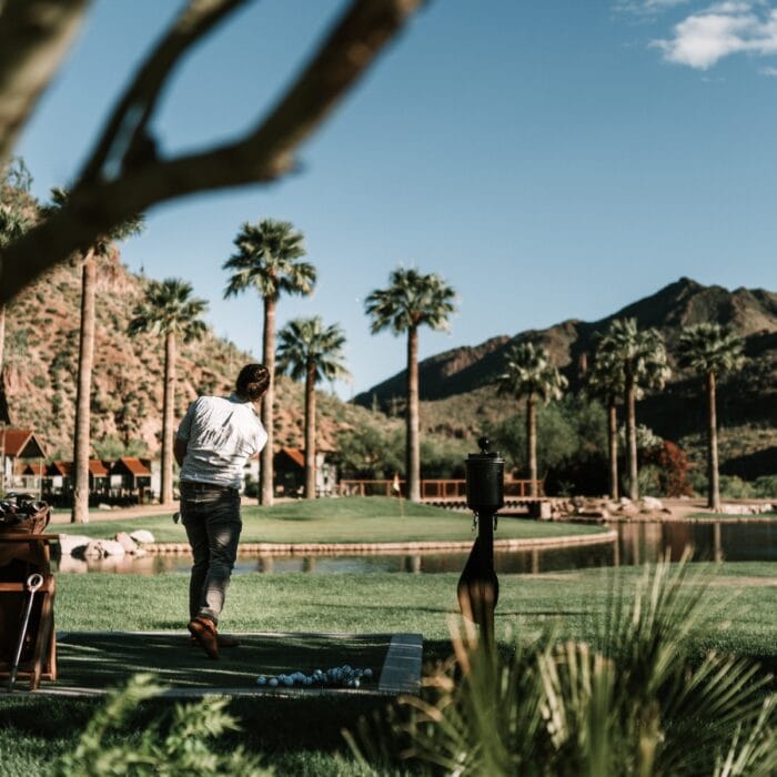 A person in the best golf apparel for Atlanta weather swings a golf club on a scenic course surrounded by palm trees, mountains, and a pond under a clear blue sky.