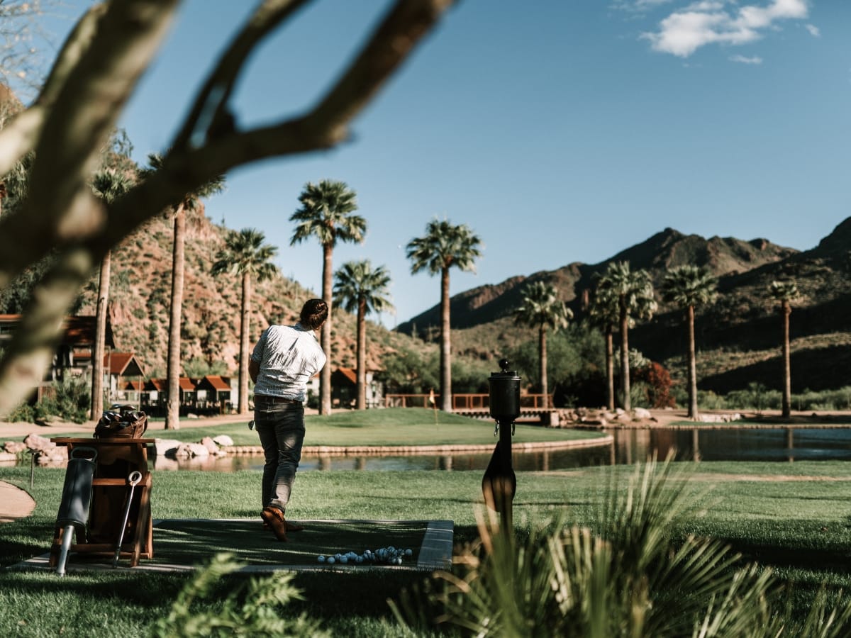 A person in the best golf apparel for Atlanta weather swings a golf club on a scenic course surrounded by palm trees, mountains, and a pond under a clear blue sky.