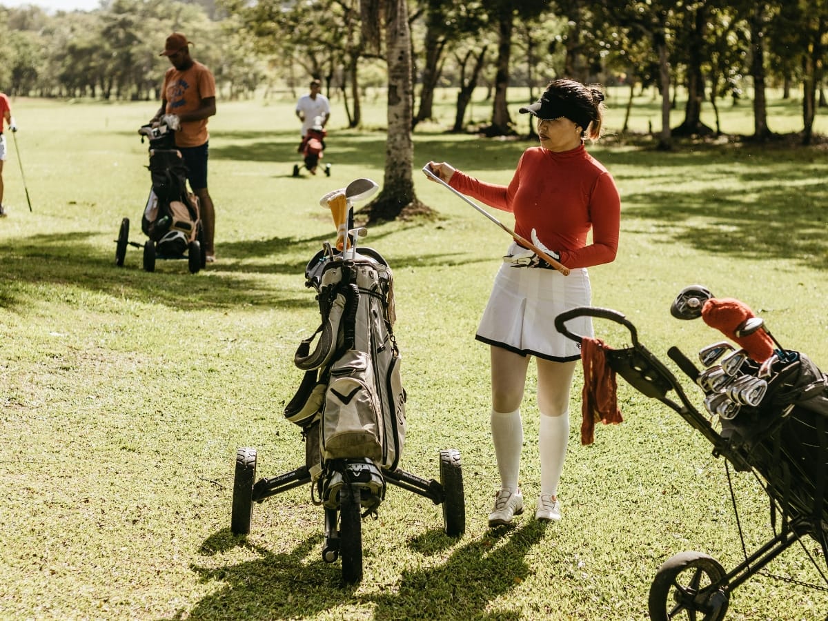 A woman in a red top and white skirt from popular golf clothing brands stands near a golf bag, reading a paper on a golf course with other players in the background.