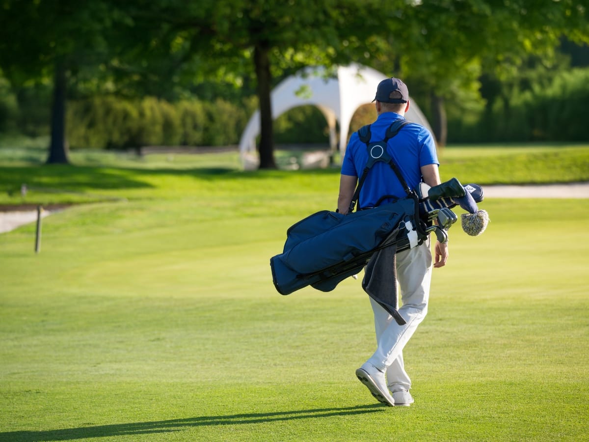 A man in a blue shirt and white pants—styled from popular golf clothing brands—carries a golf bag while walking on a golf course with green grass and trees in the background.