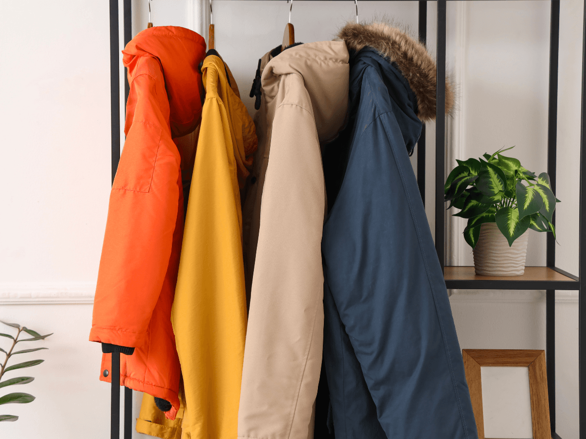 Four winter jackets in orange, yellow, beige, and blue with faux fur hoods hang on a metal rack next to a potted plant—perfect for staying cozy while keeping up with the latest Golf Apparel Trends.