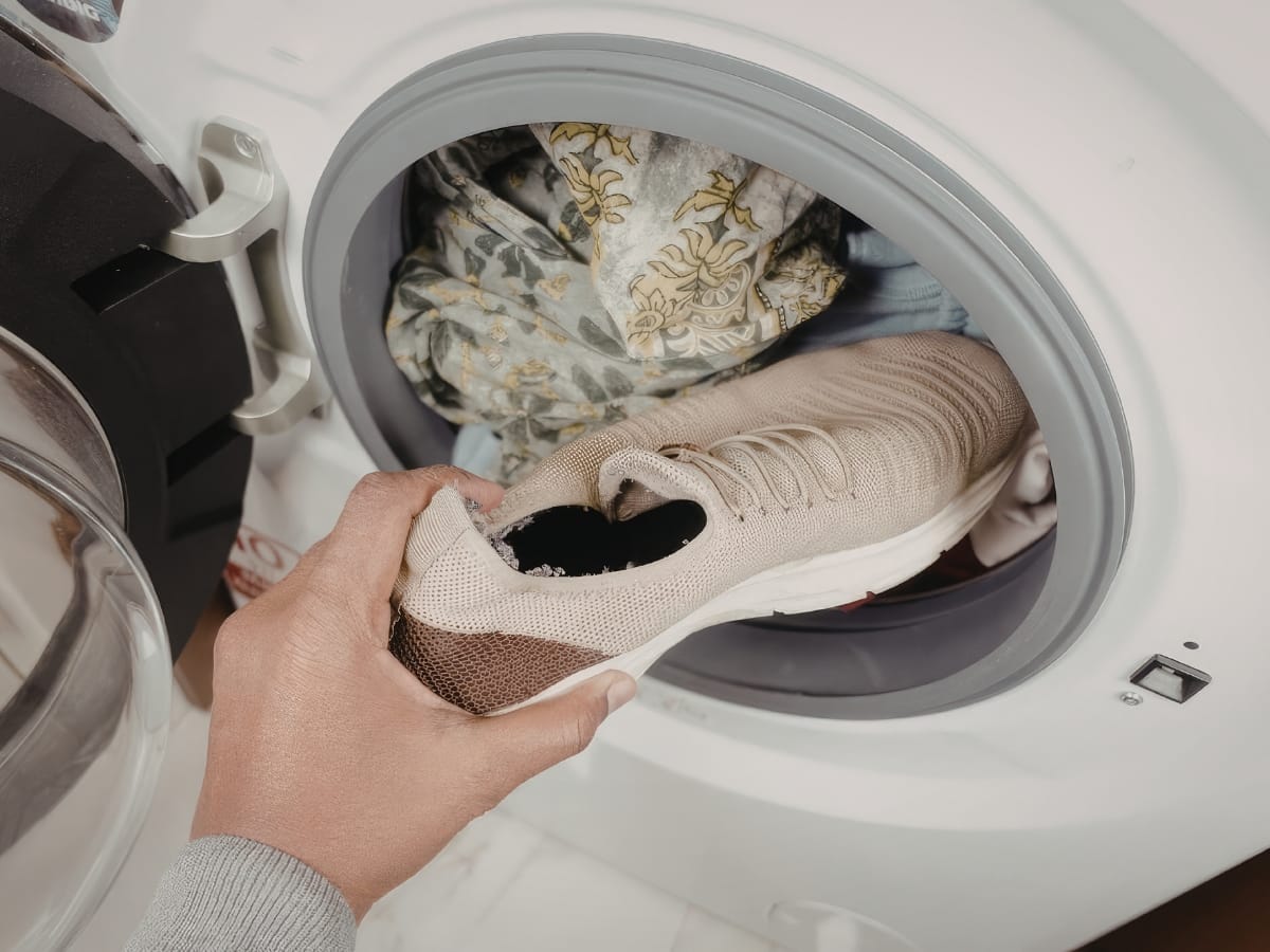 A hand is placing a beige sneaker into a washing machine filled with patterned laundry, preparing it alongside Rainy Weather Golf Gear for the next game.