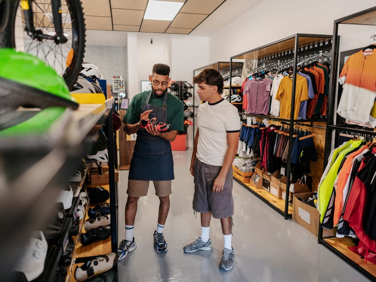 A store employee shows a cycling shoe to a customer in a sports shop with racks of clothing, gear, and even the best golf shoes on display.