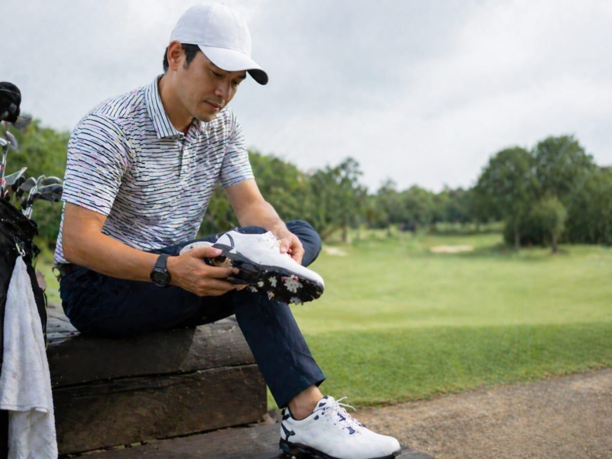 A man in a cap sits on a bench at a golf course, examining the sole of his best golf shoes, with golf clubs and a towel nearby.