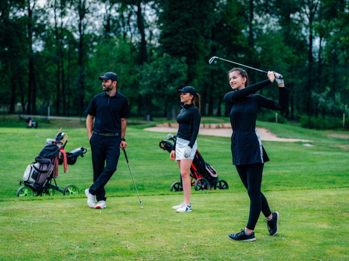 Three people stand on a golf course; two watch while one woman, dressed in stylish women's golf apparel, swings a golf club. Golf bags and trees are in the background.