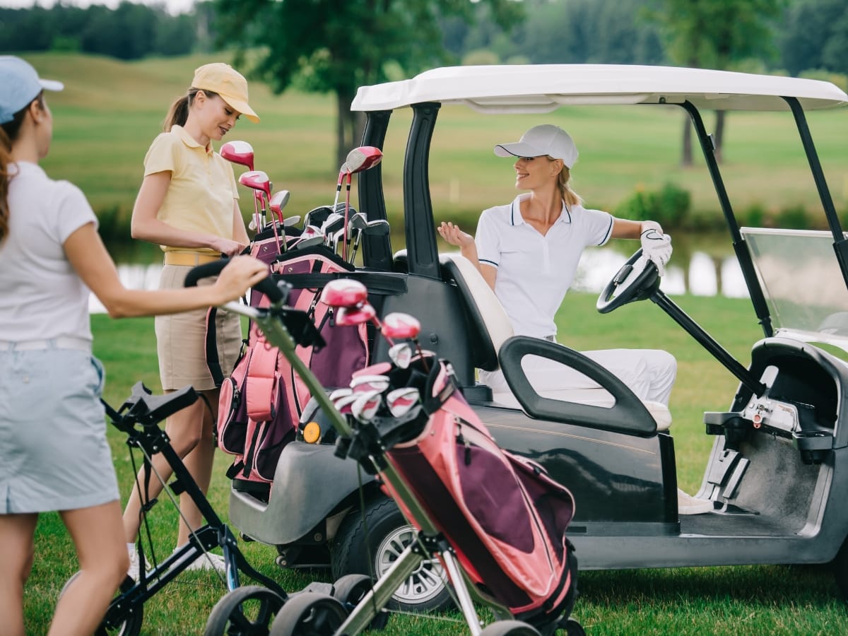 Three women in stylish women's golf apparel stand by a golf cart on a grassy course, one sitting in the driver’s seat while the others organize their clubs.