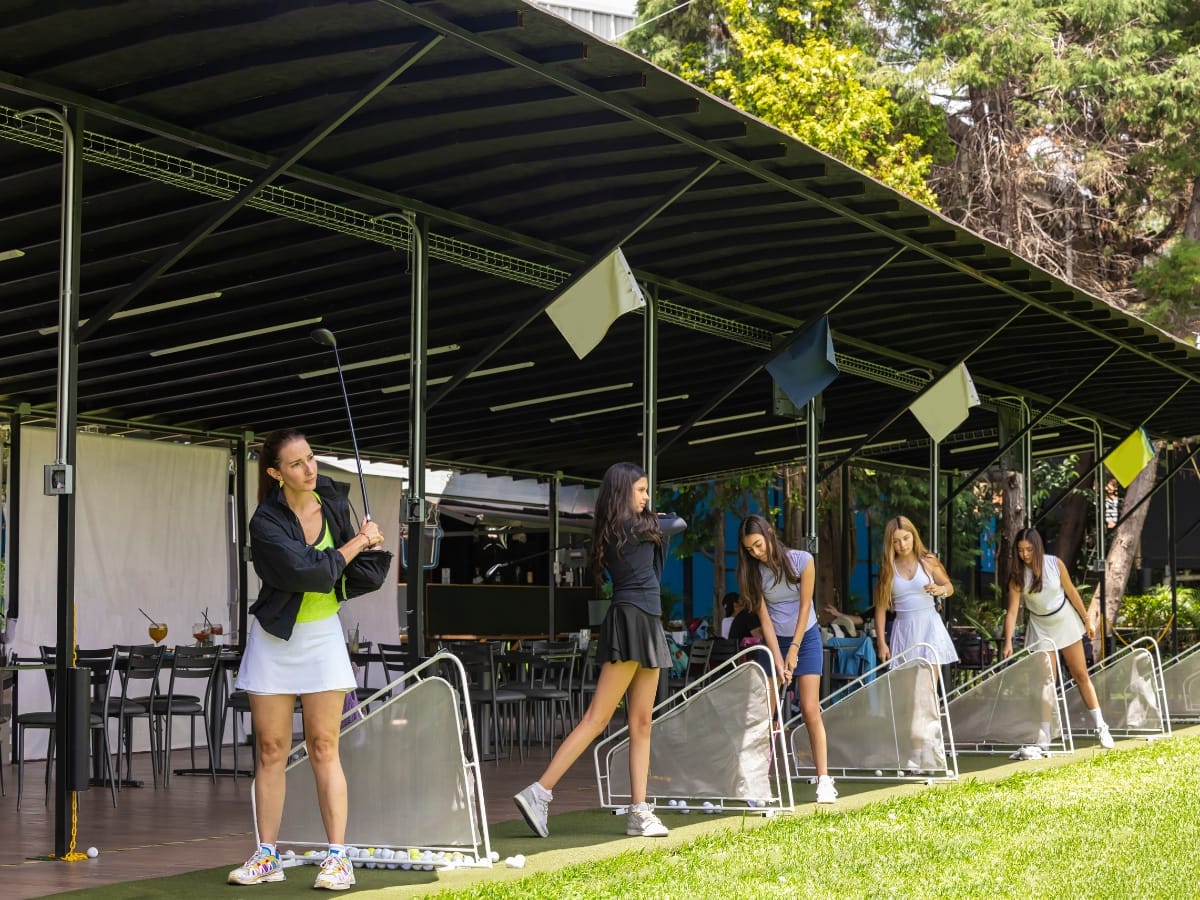 Five women in stylish women's golf apparel practice their swings at an outdoor driving range under a covered area with tables and chairs in the background.