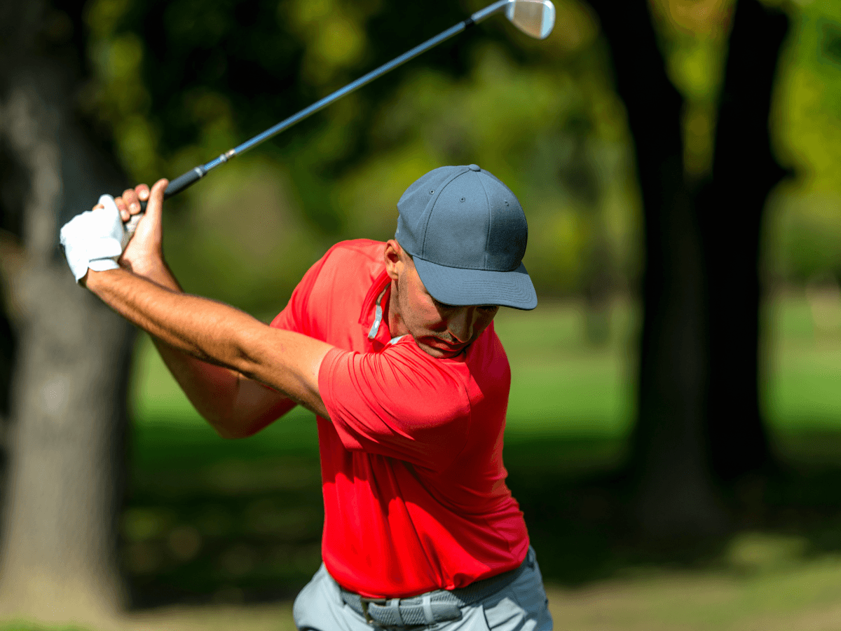 A man in a red shirt and gray cap prepares to swing a golf club on a sunny day at a golf course.