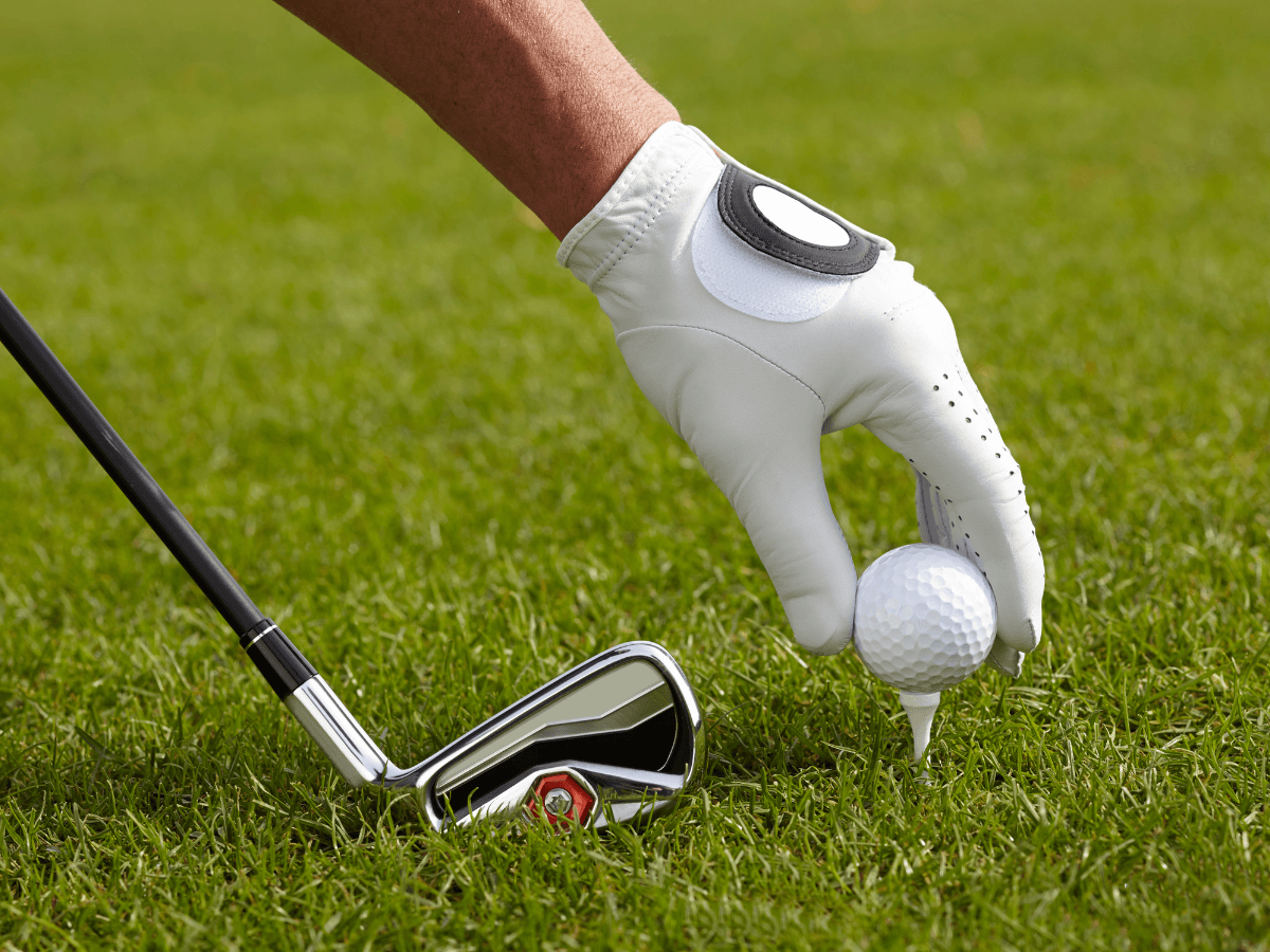 A gloved hand places a golf ball on a tee next to an iron golf club on green grass, capturing the anticipation many first time golfers feel before their opening swing.