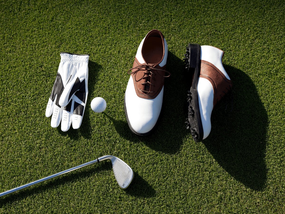 Golf shoes, a glove, a club, and a golf ball are arranged on green grass in sunlight—an inviting setup for first time golfers.