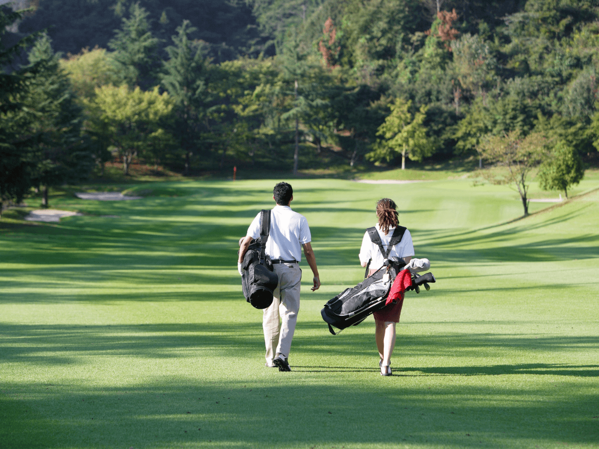 Two first time golfers carrying golf bags walk side by side on a grassy golf course surrounded by trees on a sunny day.