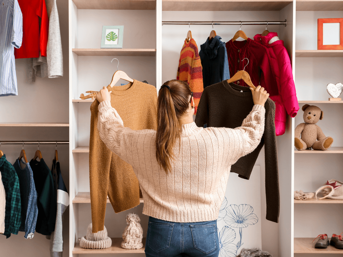 A woman stands in front of an open wardrobe, holding up two sweaters for comparison, with clothes, golf apparel, toys, and decor items visible on the shelves.