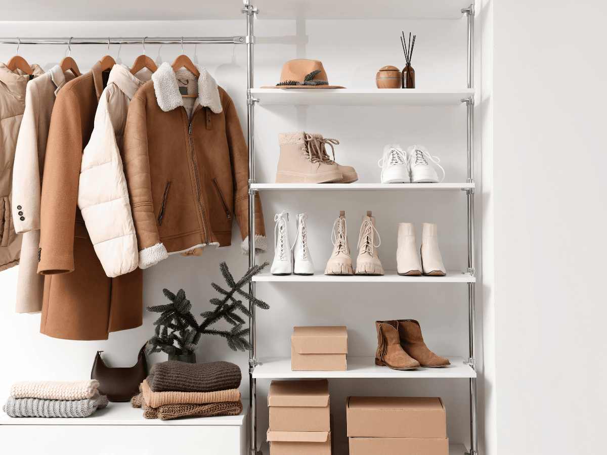 A minimalist closet features tan and beige coats on hangers, folded sweaters, golf apparel, boots, shoes, shoe boxes, a hat, and decorative items arranged on shelves.