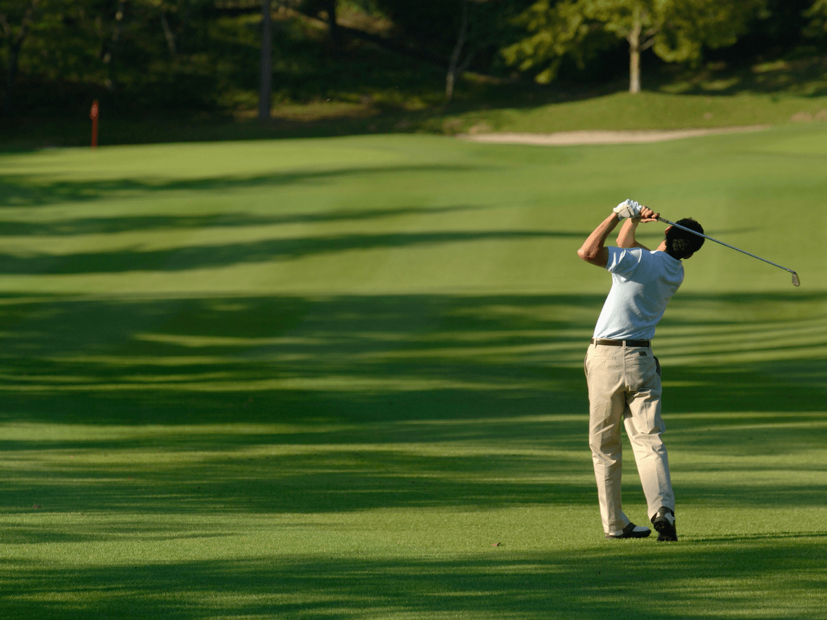 A golfer on a green fairway, wearing the best golf shoes, watches his shot after swinging a golf club, surrounded by trees and sunlight.