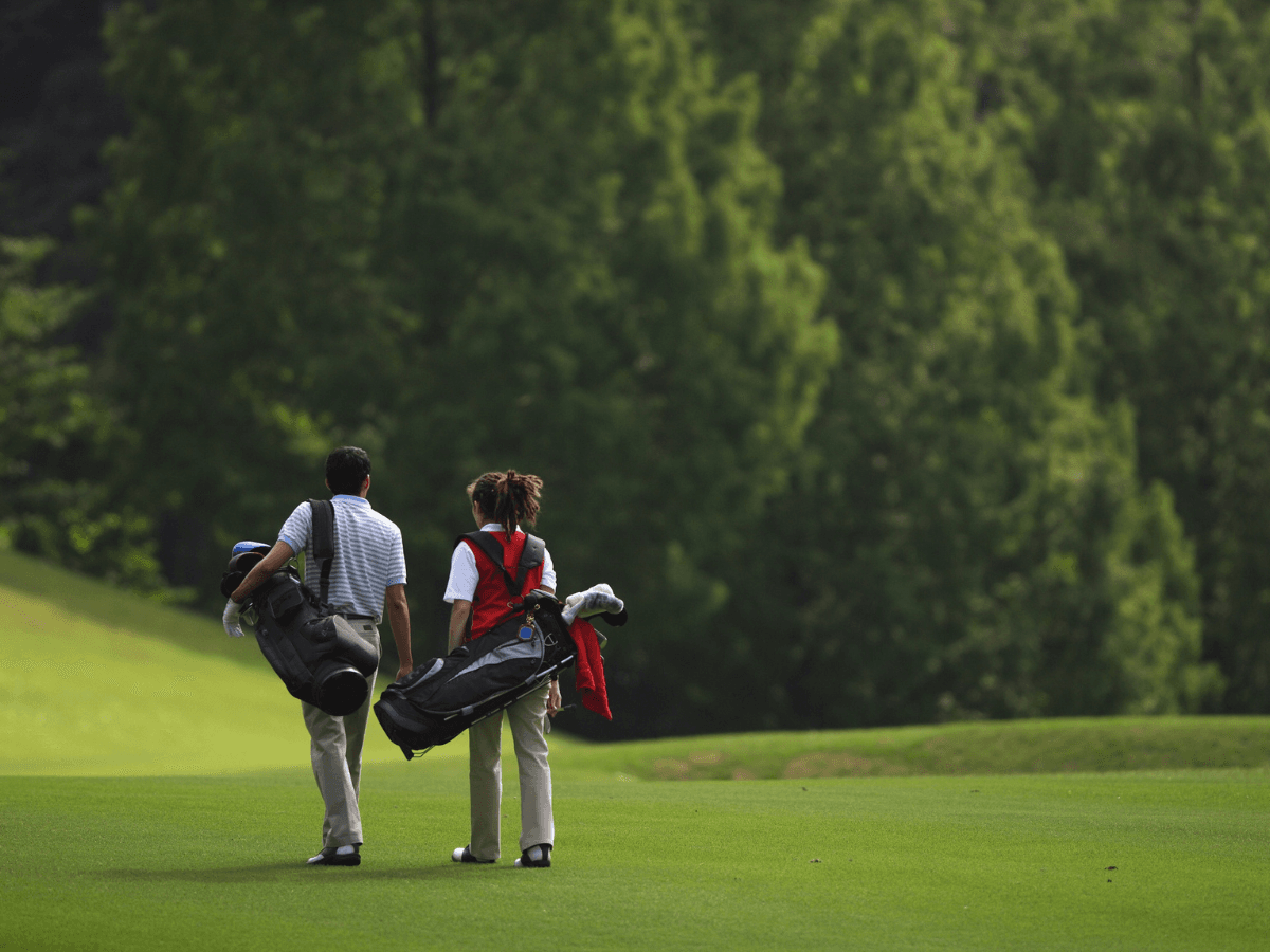 Two people carrying golf bags, dressed in trendy golf outfits, walk across a green golf course with trees in the background.
