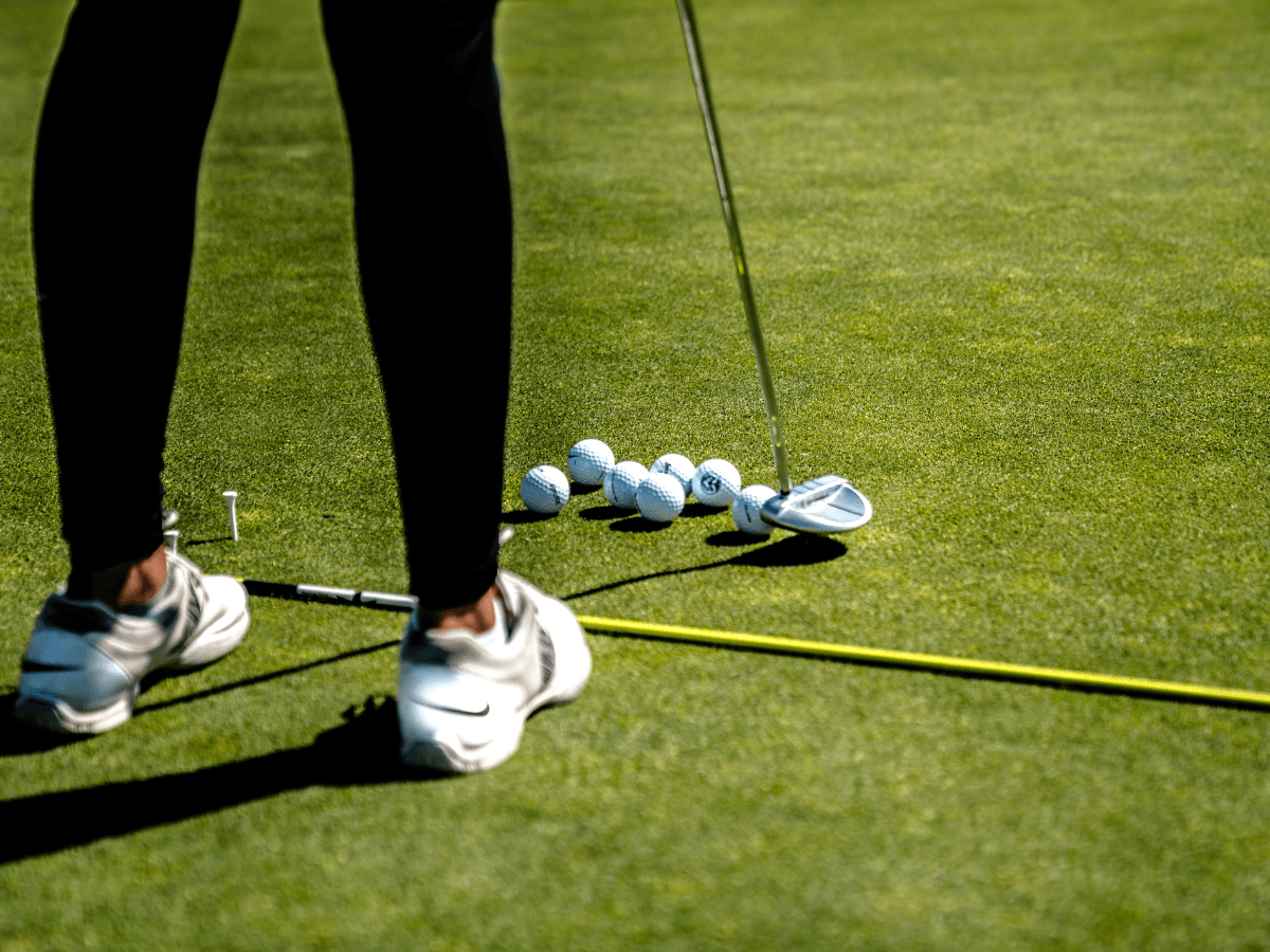 Person practicing putting on a golf green with multiple golf balls, a putter, and alignment stick visible on the grass while wearing waterproof golf shoes.