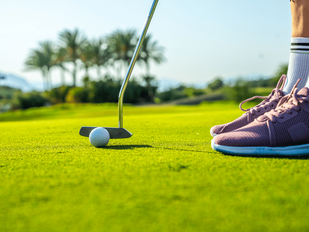 A person in pink waterproof golf shoes prepares to putt a golf ball on a green golf course, with palm trees in the background.