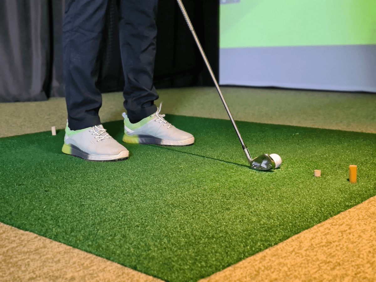 Person wearing white waterproof golf shoes prepares to hit a golf ball with an iron club on an indoor putting green. Golf tees are scattered nearby.