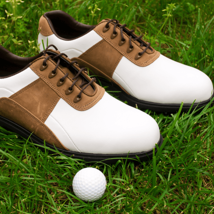 A pair of affordable golf shoes for men in brown and white, complete with brown laces, are pictured on grass next to a white golf ball.