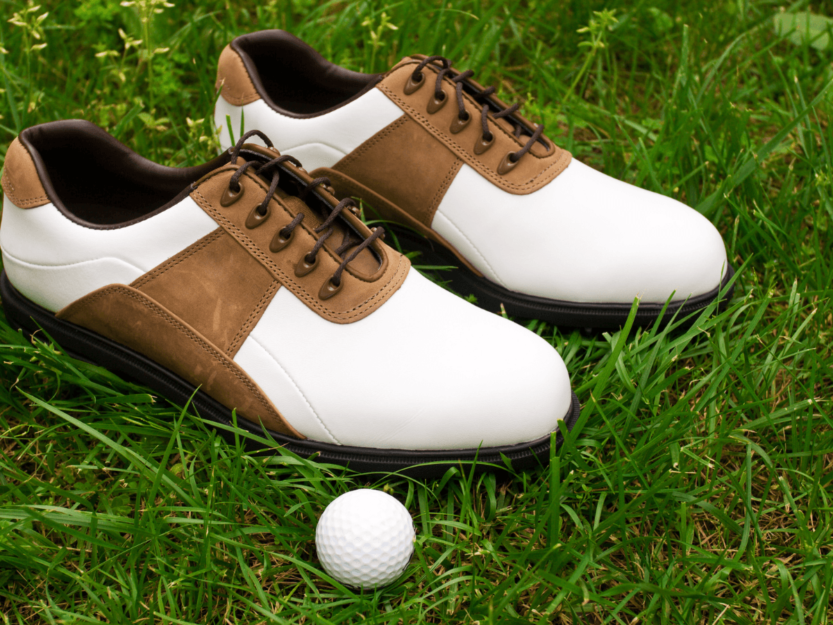 A pair of affordable golf shoes for men in brown and white, complete with brown laces, are pictured on grass next to a white golf ball.