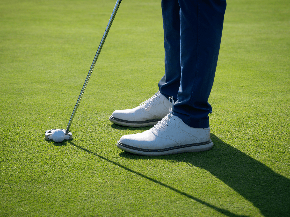 Person wearing affordable golf shoes for men and blue pants stands on a golf green, preparing to putt a golf ball with a putter.