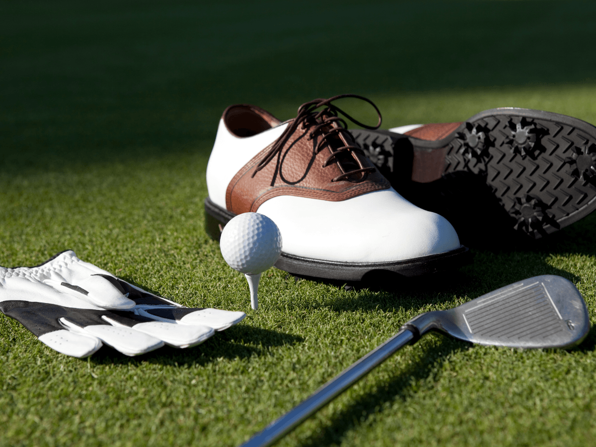 A golf glove, affordable golf shoes for men, a golf ball on a tee, and a golf club are arranged on grass.