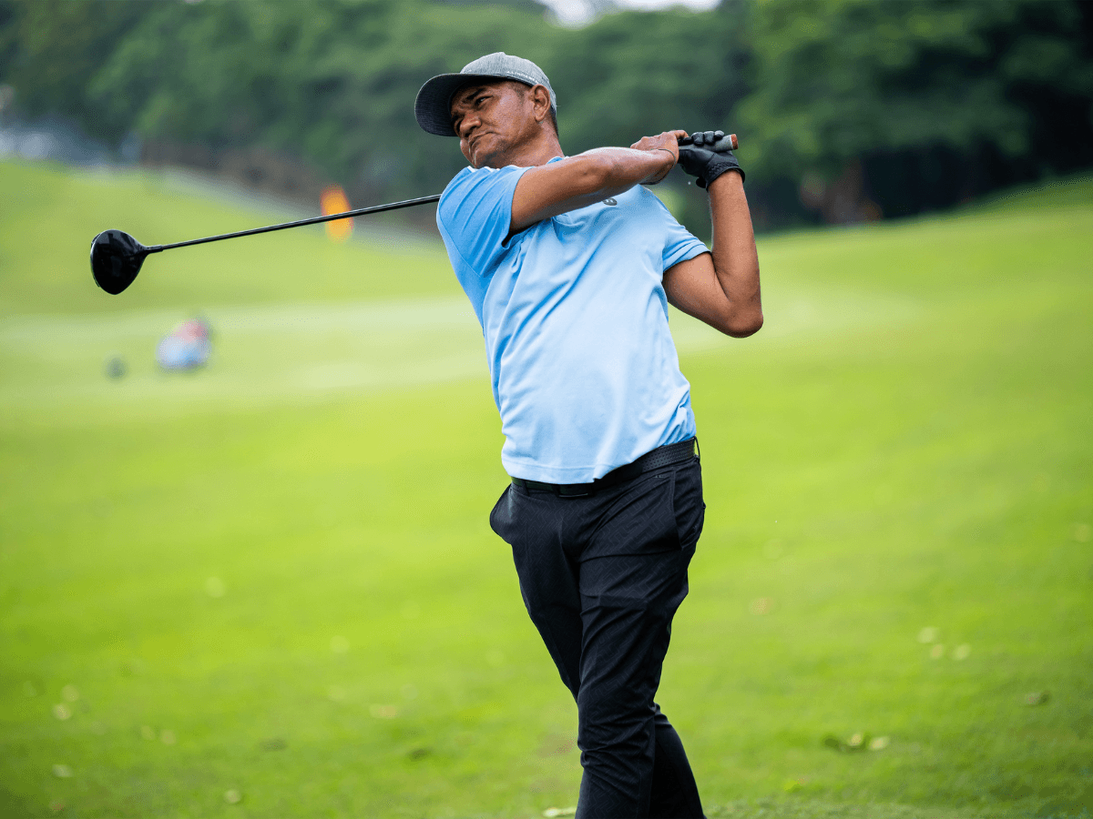 A man in a light blue shirt and cap from one of the top eco friendly golf apparel brands completes a golf swing on a green course, with trees and blurred background visible.