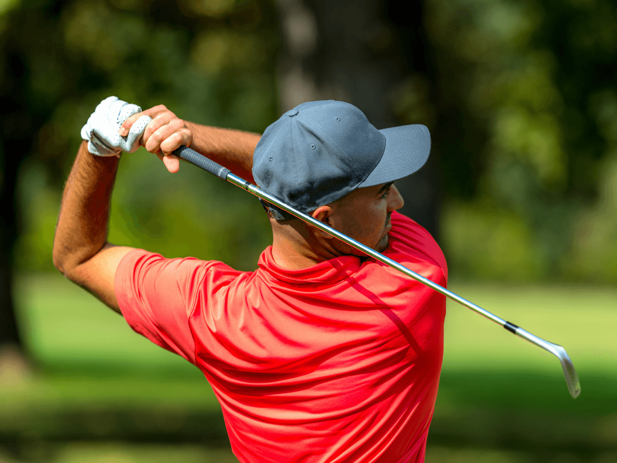 A man in a red shirt and gray cap—both from eco friendly golf apparel brands—swings a golf club on a green golf course, viewed from behind.